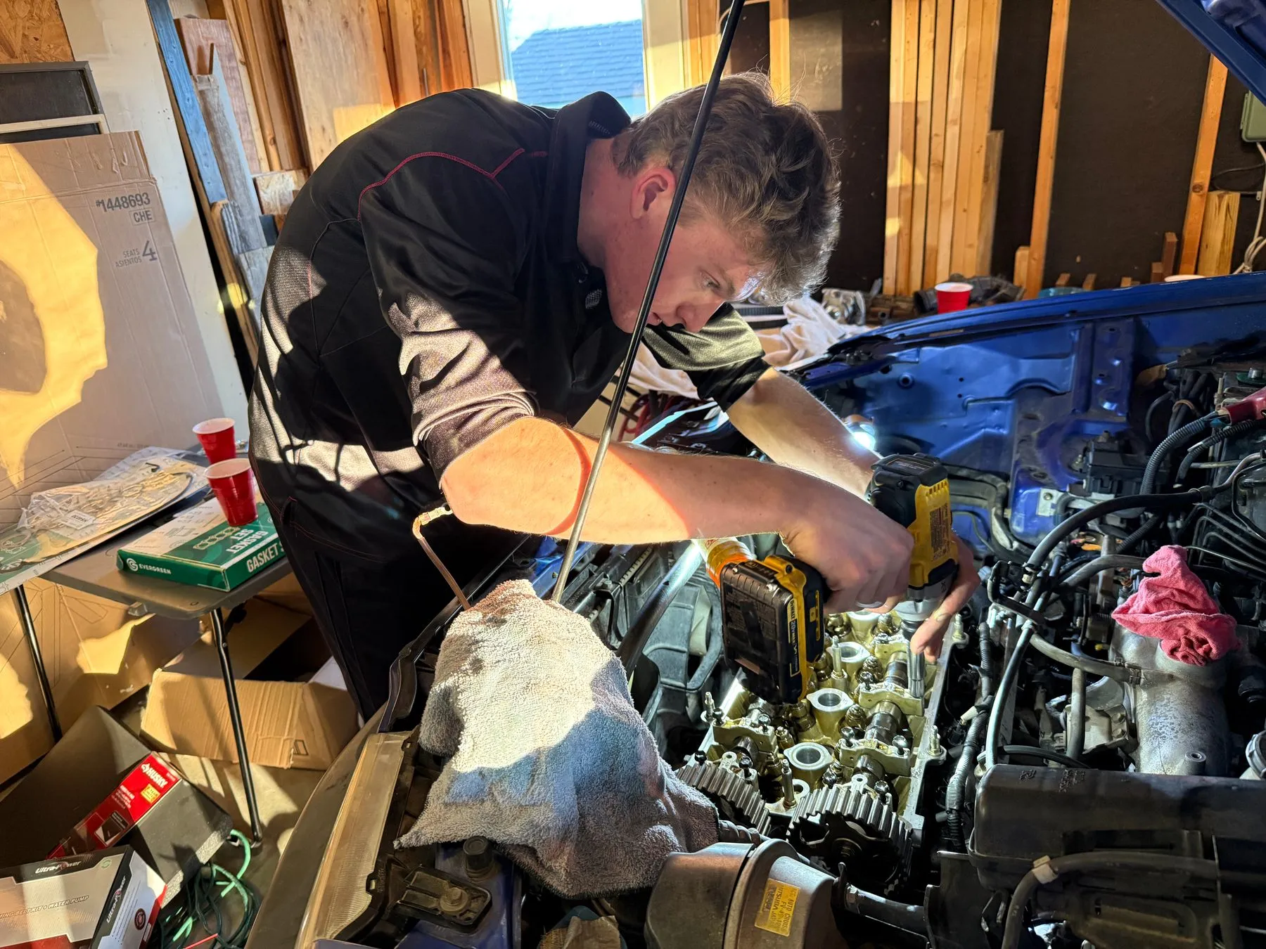 Leaning over the open engine bay of a blue car, using a cordless driver on the valve cover bolts