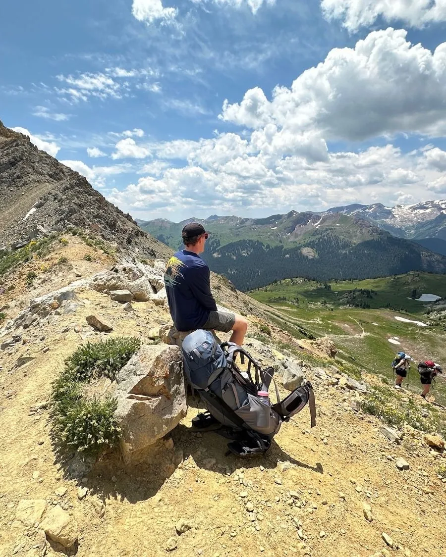 Resting on a rocky ridge, looking out over an alpine valley while two hikers climb the trail below