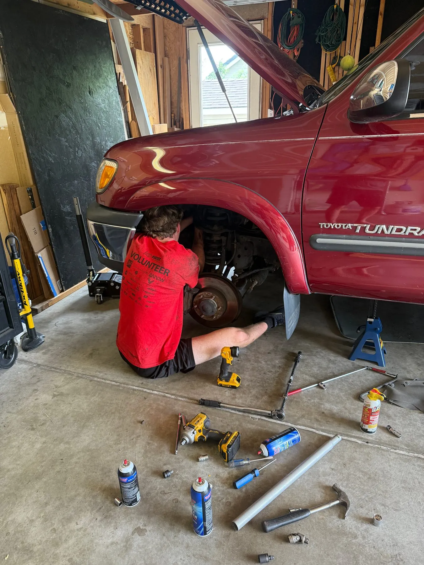 Sitting on a garage floor beside a red Toyota Tundra, working on the front brake assembly with tools spread around
