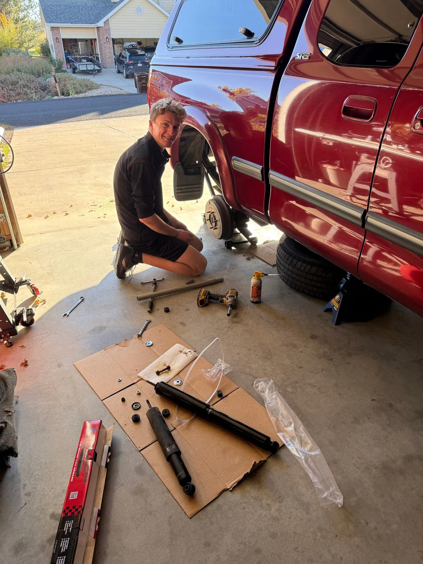 Kneeling beside a red pickup in the driveway, smiling, with the old and new shock absorbers laid out on cardboard
